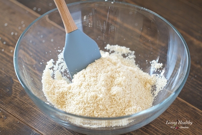 close up of a glass bowl of almond flour with spatula sitting on wooden table